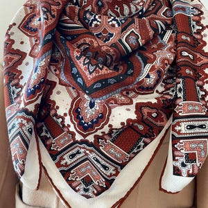May include: A square scarf with a detailed paisley pattern in shades of red, white, navy blue, and black. The scarf has a white border and is draped over a neutral-colored garment.