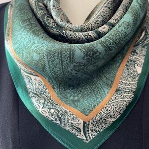 May include: A square silk scarf in shades of green, with a detailed paisley pattern. The scarf has a dark green border with a thin gold-coloured trim. The scarf is draped, showcasing its design and texture.