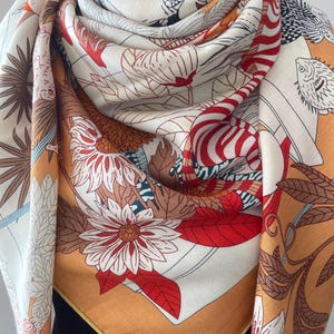 Puede incluir: Un pañuelo cuadrado con un estampado vibrante de flores, hojas y peces estilizados en tonos naranja, rojo, blanco y marrón. La tela tiene una textura visible de espiga y está drapeada para mostrar el intrincado patrón botánico y acuático.