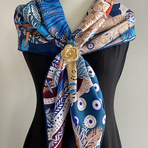 May include: A blue, orange and white patterned silk scarf with a gold floral brooch. The scarf is tied around the neck of a mannequin wearing a black dress.