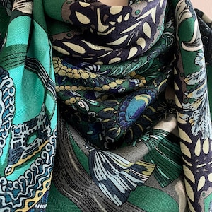 Peut inclure: &Eacute;charpe &agrave; motifs verts et bleus, avec des motifs floraux et abstraits. L'&eacute;charpe pr&eacute;sente des couleurs sarcelle, marine et vert olive. Le tissu semble doux et fluide, avec un tissage en chevrons subtil. L'&eacute;charpe est pli&eacute;e.