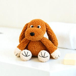 May include: A handmade brown crochet dog plush toy with white accents on the paws. The dog has a black nose, large eyes, and long floppy ears. The plush is sitting on a white surface.