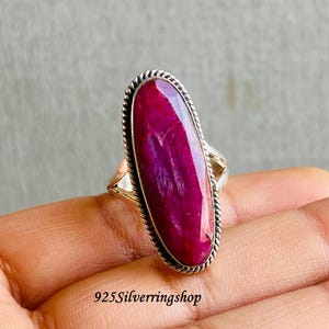 May include: A silver ring with a large, oval-shaped, deep pink gemstone. The gemstone is set in a silver band with a twisted rope design around the edge. The ring is displayed on a hand.