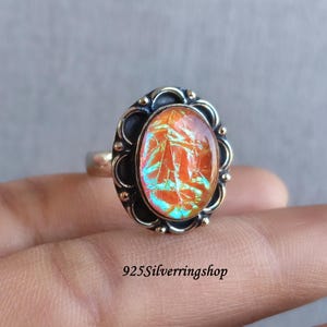May include: A silver ring with an oval-shaped, iridescent orange and blue stone. The stone is set in a dark metal frame with a scalloped edge and small gold-colored accents. The ring band is silver. The text "925Silverringshop" is visible.