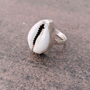 Op de afbeelding: Een zilveren ring met een witte kauri schelp in het midden. De ring ligt op een bruin, getextureerd oppervlak.