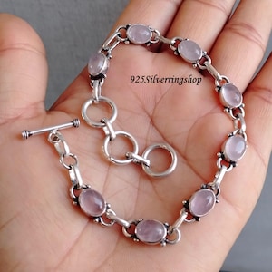 May include: A silver bracelet with a toggle clasp and oval-shaped rose quartz stones. The bracelet is on a hand and the text "925Silverringshop" is visible.