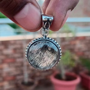 Könnte beinhalten: Silberner Anhänger mit einem runden, klaren Quarzstein mit schwarzen Einschlüssen. Der Stein ist in einer silbernen Fassung mit einem seilartigen Rand gefasst.