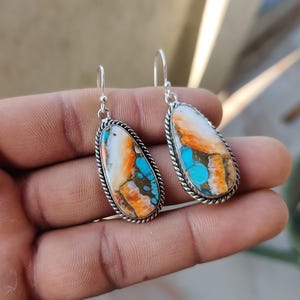 May include: A pair of silver dangle earrings with turquoise and orange stone accents. The stones are oval shaped and have a unique, marbled pattern.