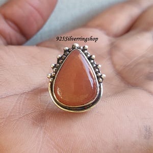 May include: A teardrop-shaped ring with a light brown sunstone set in a silver setting. The ring has a beaded design around the stone.