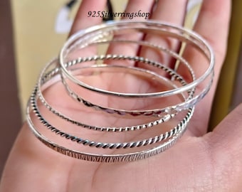 May include: A set of five silver bangles with different textures, including a hammered finish, a twisted design, and a beaded edge. The bangles are stacked on a hand.