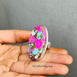 May include: A silver ring featuring a large, oval-shaped stone with a vibrant pattern. The stone showcases a mix of colors, including bright pink, light blue, white, and brown. The ring's band is a simple, polished silver.