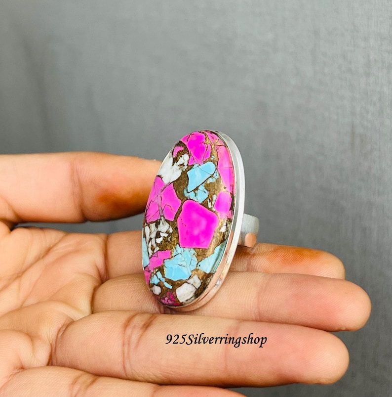 May include: A silver ring featuring a large, oval-shaped stone with a vibrant pattern. The stone showcases a mix of colors, including bright pink, light blue, white, and brown. The ring's band is a simple, polished silver.