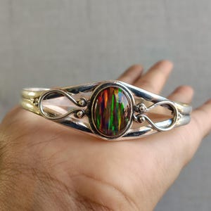 May include: A silver cuff bracelet featuring an oval, iridescent opal stone. The opal displays a vibrant array of colors, including green, red, and yellow. The bracelet has a decorative, openwork design with curved lines and small, round accents.