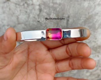 May include: A silver bangle bracelet with a large, rectangular, pink and orange gemstone set in the center. The bracelet is on a person's hand.