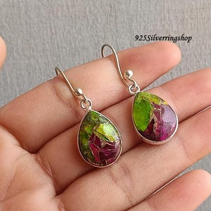 May include: A pair of teardrop-shaped earrings with a silver-colored frame. The earrings feature a vibrant mix of green, gold, and deep red colors. The earrings are displayed against a neutral background.
