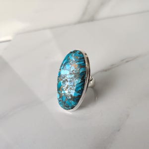 May include: A silver ring featuring a large, oval-shaped turquoise stone with a mosaic pattern of blue, gold, and white. The stone is set in a polished silver band.