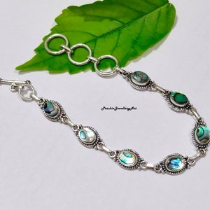 Puede incluir: Pulsera de plata con cierre de palanca y detalles ovalados de concha de abulón. La pulsera está sobre una superficie blanca con una hoja verde en el fondo.