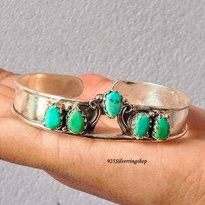 May include: A silver cuff bracelet featuring five teardrop-shaped turquoise stones set in a row. The center stone is elevated, flanked by two stones on each side. The metal band has a smooth, polished finish. The text 925Silverringshop is visible.