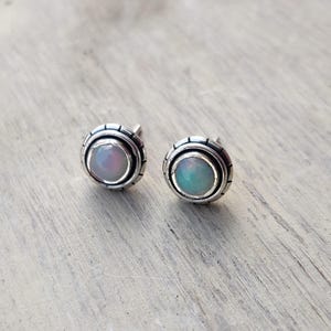 May include: A pair of silver stud earrings with a round opal gemstone in the center. The earrings have a textured silver border.