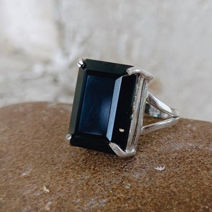 Pode incluir: Um anel de prata com uma grande pedra preta retangular. O anel tem um design simples e elegante.