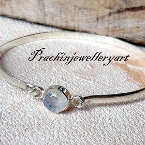 May include: A silver bangle bracelet featuring a round, white moonstone gemstone. The bracelet is open and has a simple design. The text "Prachinjewelleryart" is visible in the image.