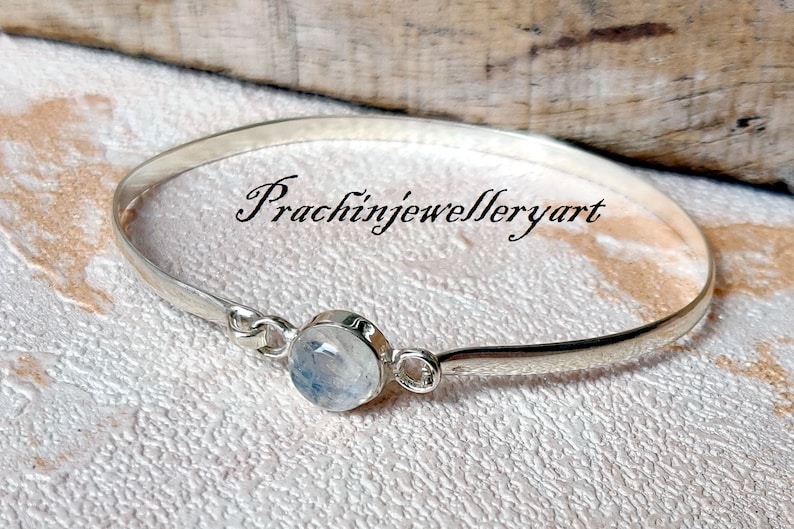 May include: A silver bangle bracelet featuring a round, white moonstone gemstone. The bracelet is open and has a simple design. The text "Prachinjewelleryart" is visible in the image.
