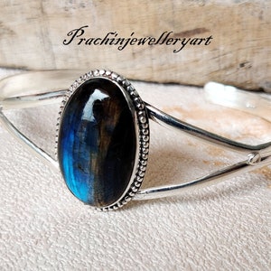 Op de afbeelding: Een zilveren armband met een grote ovale labradorit-edelsteen, gezet in een zilveren bezel. De edelsteen heeft een blauwe en zwarte iriserende glans.