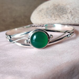 Può includere: Un braccialetto rigido in argento con una pietra preziosa verde incastonata in una lunetta rotonda. Il braccialetto ha un design delicato e a spirale.