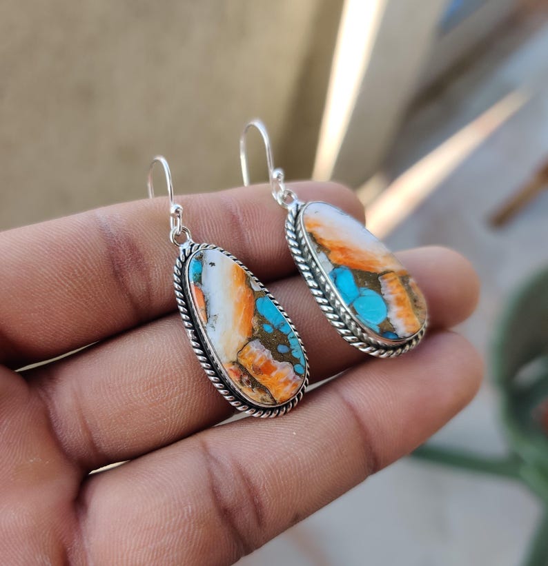 May include: A pair of silver dangle earrings with oval-shaped turquoise stones. The stones are a mix of white, orange, and blue.