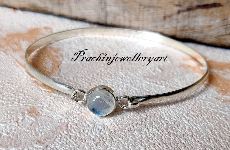 May include: A silver bangle bracelet featuring a round moonstone charm. The bracelet is open and has a simple design. The moonstone is a light blue colour with a shimmery effect.