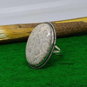 Pode incluir: Um anel de formato oval com uma grande pedra de coral fossilizado bege, apresentando um padrão natural detalhado semelhante a uma flor. A pedra está cravada numa moldura de prata polida com uma borda de corda torcida e aro de prata dividido.