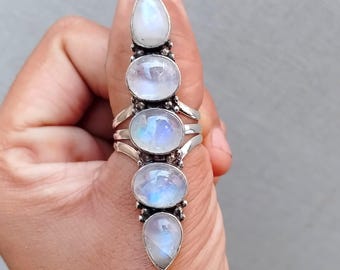 Op de afbeelding: Een lange zilveren ring met vijf iriserende maansteen cabochons. De ring heeft een langwerpig ontwerp met de stenen in een verticale rij. De maanstenen hebben een melkachtige witte en blauwe kleur. De ring wordt door een hand omhoog gehouden.