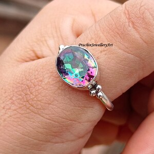 Peut inclure: Une bague en argent avec une grande pierre précieuse ovale et irisée. La pierre précieuse présente un arc-en-ciel de couleurs, notamment le rose, le vert, le bleu et le violet.
