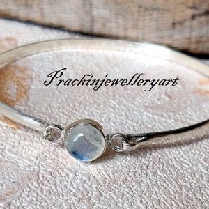 May include: A silver bangle bracelet featuring a round moonstone charm. The bracelet is open and has a simple design. The moonstone is a light blue colour with a shimmery effect.