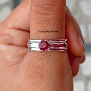May include: A silver ring with a red ruby gemstone set in the center. The ring has three bands, each with a slightly different texture.