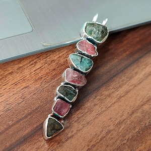 May include: A silver pendant with a row of seven faceted gemstones. The gemstones are a mix of pink, green, and black.