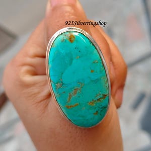 Może przedstawiać: Duży owalny pierścionek z turkusem ze srebrną oprawą. Kamień z turkusu ma brązowe i brązowe wzory macierzy. Pierścionek jest osadzony w srebrnej oprawie.