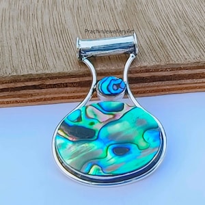 May include: A silver pendant with a round abalone shell inlay. The shell has a green, blue, and iridescent pattern. The pendant is attached to a silver bail.