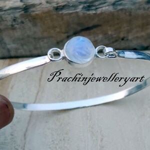 May include: A silver bangle bracelet featuring a round moonstone charm. The bracelet is open and has a simple design. The moonstone is a pale blue colour with a shimmering effect.