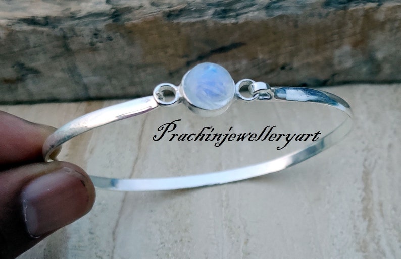 May include: A silver bangle bracelet featuring a round moonstone charm. The bracelet is open and has a simple design. The moonstone is a pale blue colour with a shimmering effect.