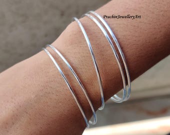 May include: Five silver bangles stacked on a person's wrist. The bangles are thin and have a smooth, polished finish.