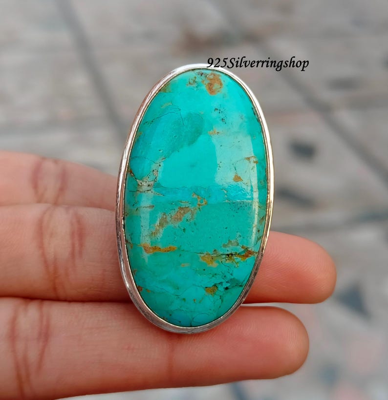 May include: A large, oval turquoise gemstone ring with a silver band. The turquoise stone is a vibrant blue-green color with brown veining. The ring is held in a hand, and the text "925Silverringshop" is visible.