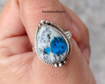 May include: A silver ring featuring a teardrop-shaped stone with a white, gray, and blue pattern. The stone is set in a silver bezel with a beaded edge. The ring is held by a hand.
