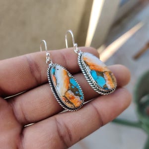 May include: A pair of silver dangle earrings with oval-shaped turquoise stones. The stones are a mix of white, orange, and blue.