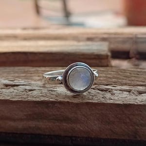 May include: A silver ring with a round moonstone set in a bezel setting. The ring has a simple band with small decorative accents.
