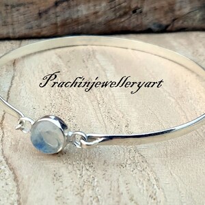 May include: A silver bangle bracelet with a round, white moonstone charm. The bracelet is on a wooden surface. The text "Prachinjewelleryart" is visible in the image.