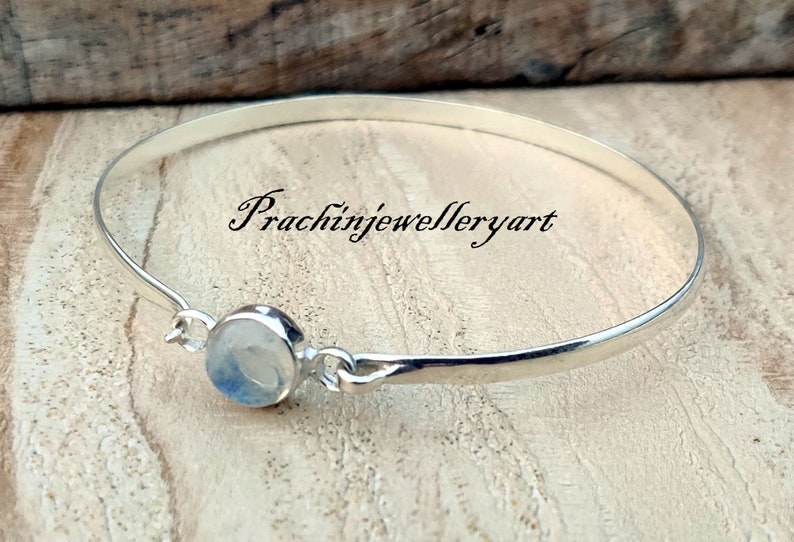 May include: A silver bangle bracelet with a round, white moonstone charm. The bracelet is on a wooden surface. The text "Prachinjewelleryart" is visible in the image.