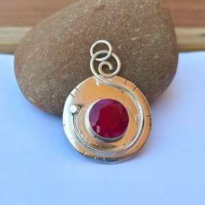 May include: A silver pendant with a red gemstone set in a circular design. The pendant has a swirl design around the gemstone and a bail for attaching a chain.