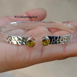 Op de afbeelding: Een zilveren gegraveerde armband met twee gele edelstenen. De armband is open en heeft een gegraveerde textuur.