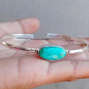 May include: A silver bangle bracelet with a turquoise oval gemstone. The bracelet is simple and elegant, with a smooth finish.